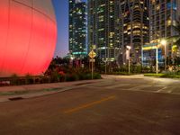 a street at dusk with a large red ball in the middle of it and many tall buildings in the background