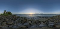 a large body of water surrounded by rocks next to a cityscape with the sun coming out