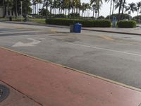 a sidewalk next to a blue garbage can on a red brick paved city street, surrounded by palm trees