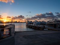 a sunrise over the ocean with many boats parked near by a dock and a city skyline in the distance