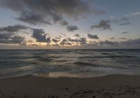 a person standing on top of a sandy beach next to the ocean at sunset with clouds in the background