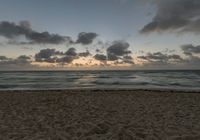 a person standing on top of a sandy beach next to the ocean at sunset with clouds in the background