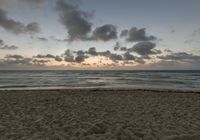 a person standing on top of a sandy beach next to the ocean at sunset with clouds in the background