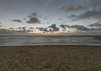 a person standing on top of a sandy beach next to the ocean at sunset with clouds in the background