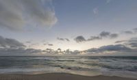 a picture of a beach with waves coming towards shore and clouds in the distance with a sunset in the background