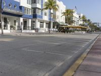 a deserted street lined with shops and umbrellas next to palm trees and white buildings