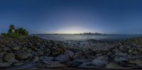 the sun is shining brightly behind the city skyline from a rocky shoreline at night time