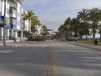 a wide empty street lined with tall buildings near palm trees and a beach area in the background