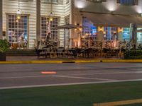 a building that has tables outside at night on a street corner, a red light is lit from a neon