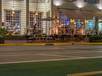 a building that has tables outside at night on a street corner, a red light is lit from a neon