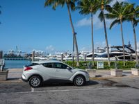 a car is parked in the lot by a dock with yachts and palm trees