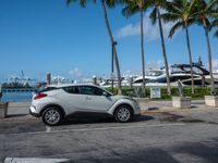 a car is parked in the lot by a dock with yachts and palm trees