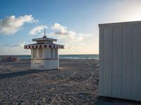 a lifeguard house is on a sandy beach near the ocean's edge with two lounge chairs in the sand and an umbrella shade