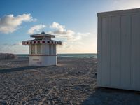 a lifeguard house is on a sandy beach near the ocean's edge with two lounge chairs in the sand and an umbrella shade