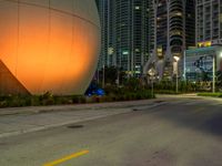 a big white sphere that is in the middle of a street at night, with skyscrapers in the background