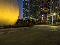 a city street with a giant yellow ball near a parking lot at night and tall buildings behind it