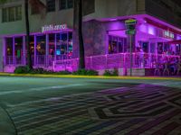 a cafe with neon lights, near the street with people sitting outside at night, and in front of a multi - colored building