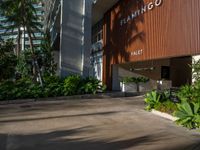 the entrance to an apartment with palms trees and bushes in the foreground, next to a concrete driveway