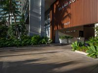 the entrance to an apartment with palms trees and bushes in the foreground, next to a concrete driveway