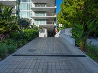 the entrance to an apartment with palms trees and bushes in the foreground, next to a concrete driveway