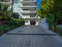 the entrance to an apartment with palms trees and bushes in the foreground, next to a concrete driveway