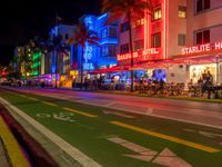 people sit outside an aldi hotel in the evening, and neon signs glow on the sidewalk