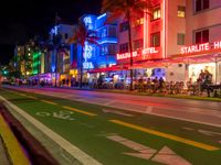 people sit outside an aldi hotel in the evening, and neon signs glow on the sidewalk