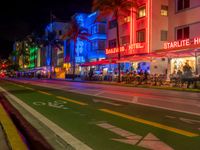 people sit outside an aldi hotel in the evening, and neon signs glow on the sidewalk