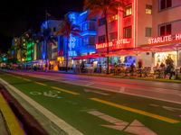 people sit outside an aldi hotel in the evening, and neon signs glow on the sidewalk