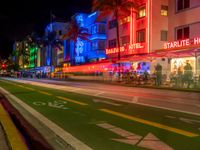 people sit outside an aldi hotel in the evening, and neon signs glow on the sidewalk