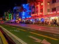 people sit outside an aldi hotel in the evening, and neon signs glow on the sidewalk