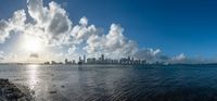 the man walking along the shore of the ocean next to the city skylines, holding a surfboard, against a blue sky