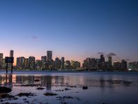 a city skyline, with water, at sunset, and rocks along the shore, as seen from the water