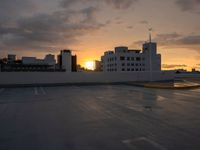 sunset over a parking lot with buildings in the background and cloudy sky above them,