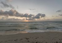 a beach with the sun setting behind clouds and waves washing up on the sand with the ocean in the background