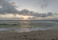 a beach with the sun setting behind clouds and waves washing up on the sand with the ocean in the background