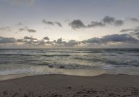 a lone surfer on a sandy beach in the ocean at sunset time, under a cloudy sky