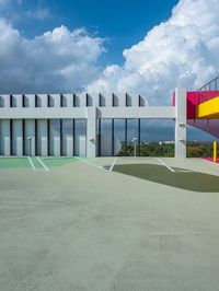 a concrete basketball court with colorful stairs leading into the top floor and side wall of a modern, two story building in the background