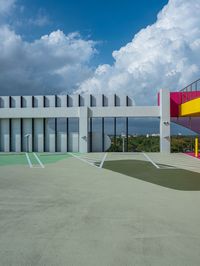 a concrete basketball court with colorful stairs leading into the top floor and side wall of a modern, two story building in the background