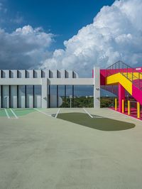 a concrete basketball court with colorful stairs leading into the top floor and side wall of a modern, two story building in the background