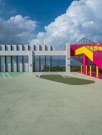 a concrete basketball court with colorful stairs leading into the top floor and side wall of a modern, two story building in the background