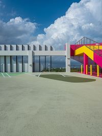 a concrete basketball court with colorful stairs leading into the top floor and side wall of a modern, two story building in the background