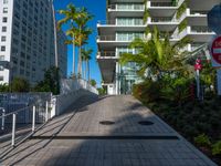 the entrance to an apartment with palms trees and bushes in the foreground, next to a concrete driveway