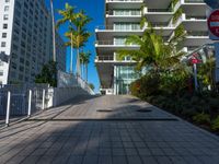 the entrance to an apartment with palms trees and bushes in the foreground, next to a concrete driveway