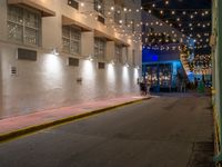 two people walking down the street in front of a building with string lights overhead from above