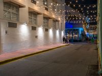 two people walking down the street in front of a building with string lights overhead from above