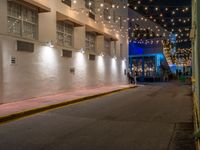 two people walking down the street in front of a building with string lights overhead from above