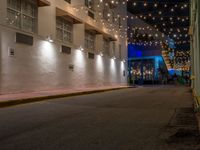 two people walking down the street in front of a building with string lights overhead from above