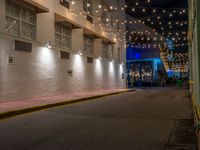 two people walking down the street in front of a building with string lights overhead from above