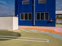colorful painted tennis court with blue building and sky behind it in the back ground, in front of it is a parking lot